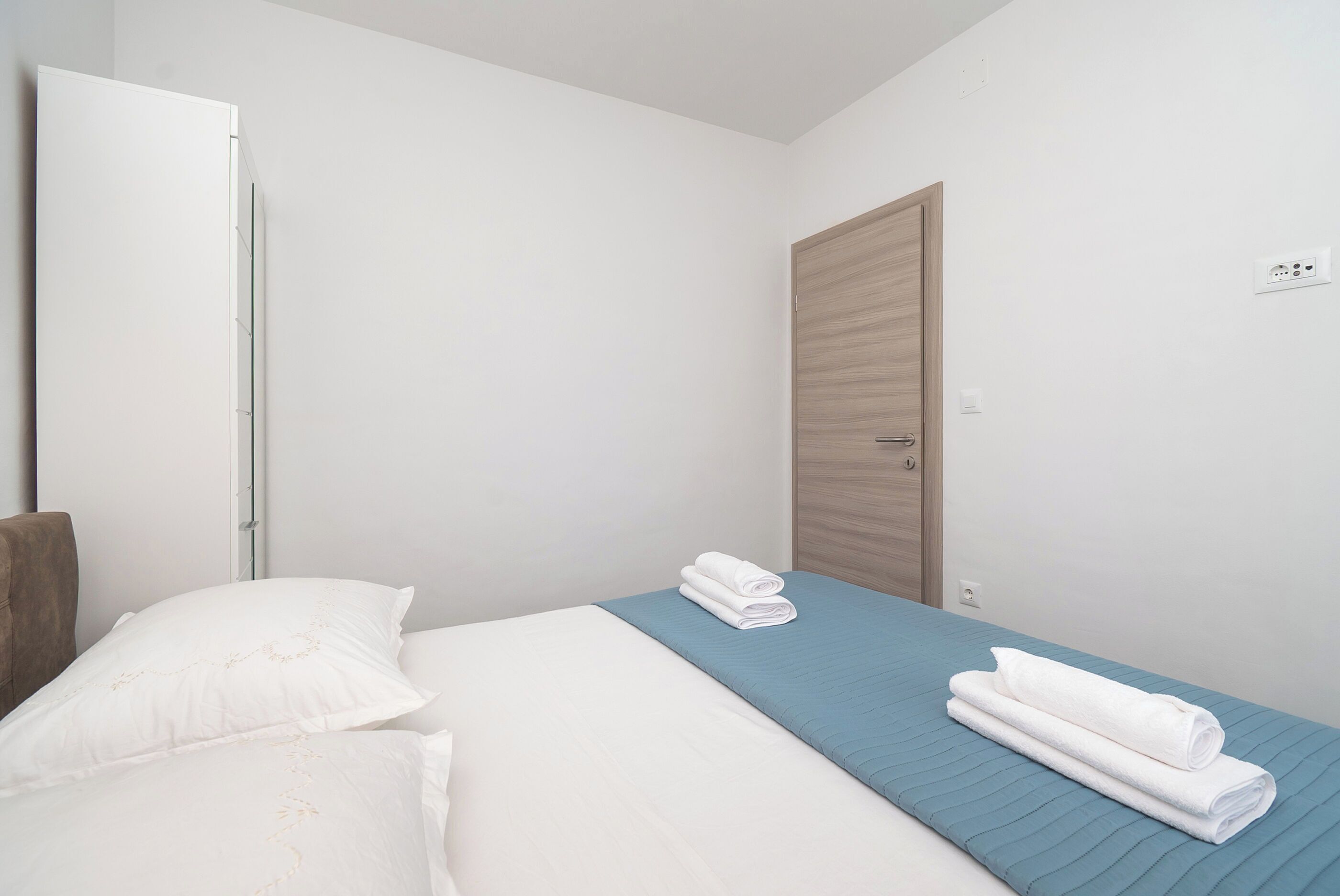 A hotel room with a bed, a blue bedspread, two rolled white towels, a wooden door, and a white wardrobe.