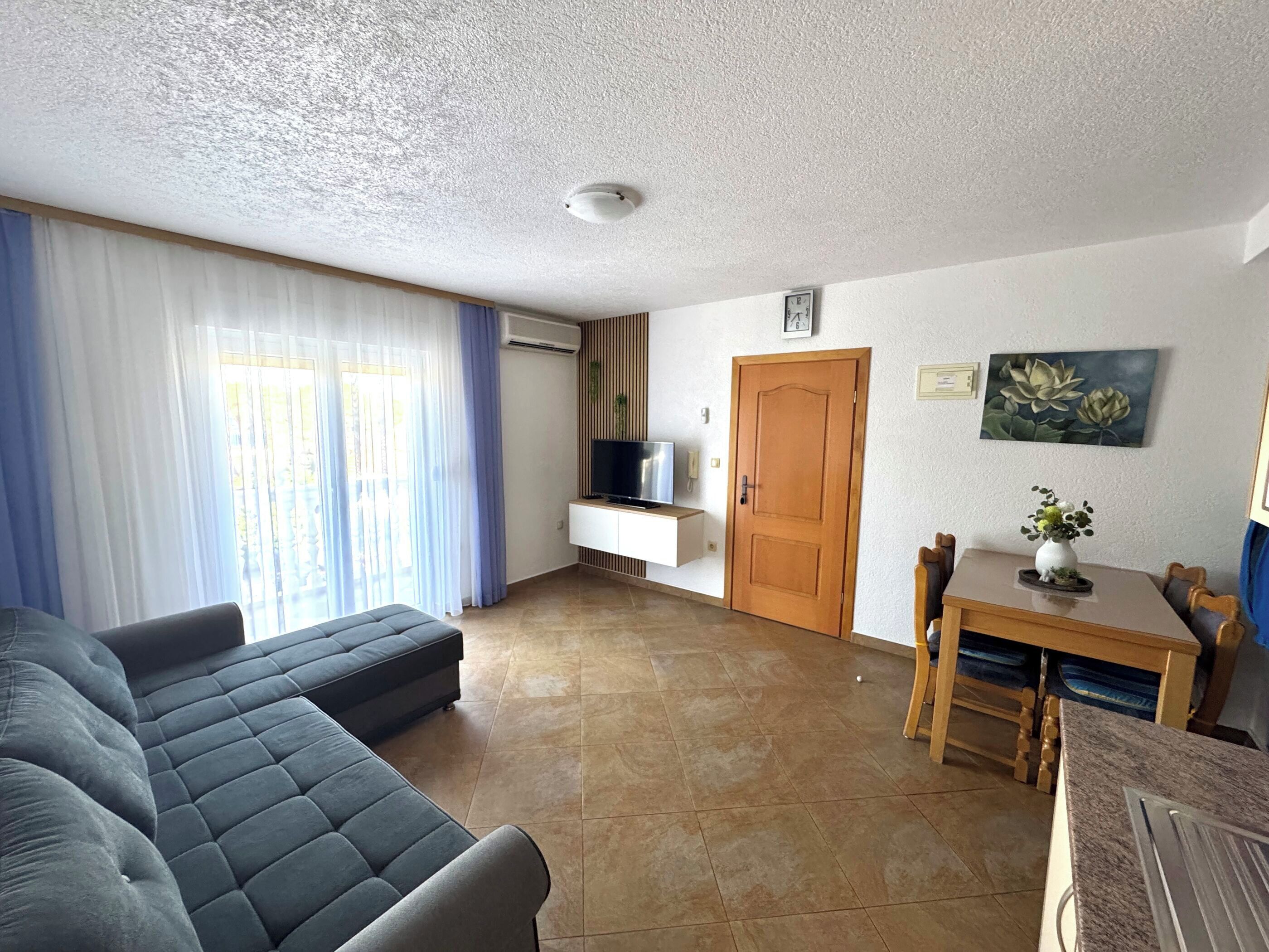 A living room with a sofa, a TV on a wall-mounted shelf, a wooden door, and a dining table with chairs.