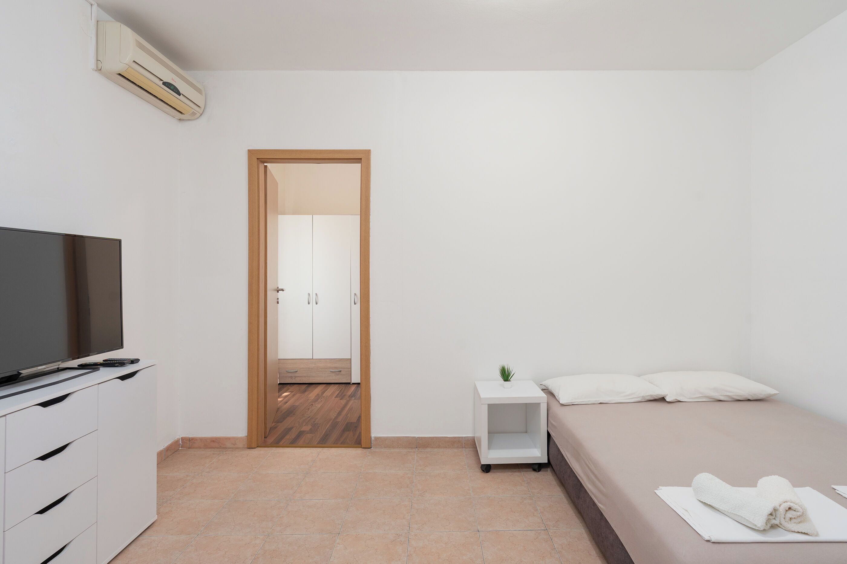 Een moderne slaapkamer met een televisie, een nachtkastje met een plant, een bed met witte kussens, en een houten deur die naar een andere kamer leidt.