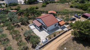 Una casa moderna de dos pisos con techo de tejas rojas, rodeada de árboles y un camino de tierra.