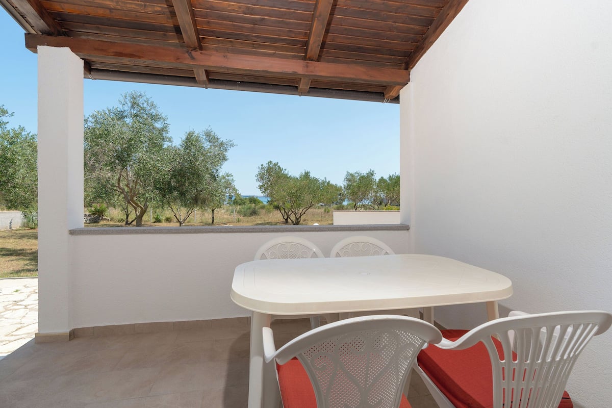 Eine Terrasse im Freien mit weißem Tisch und Stühlen, einer Holzbalkendecke und Blick auf Bäume und Himmel.