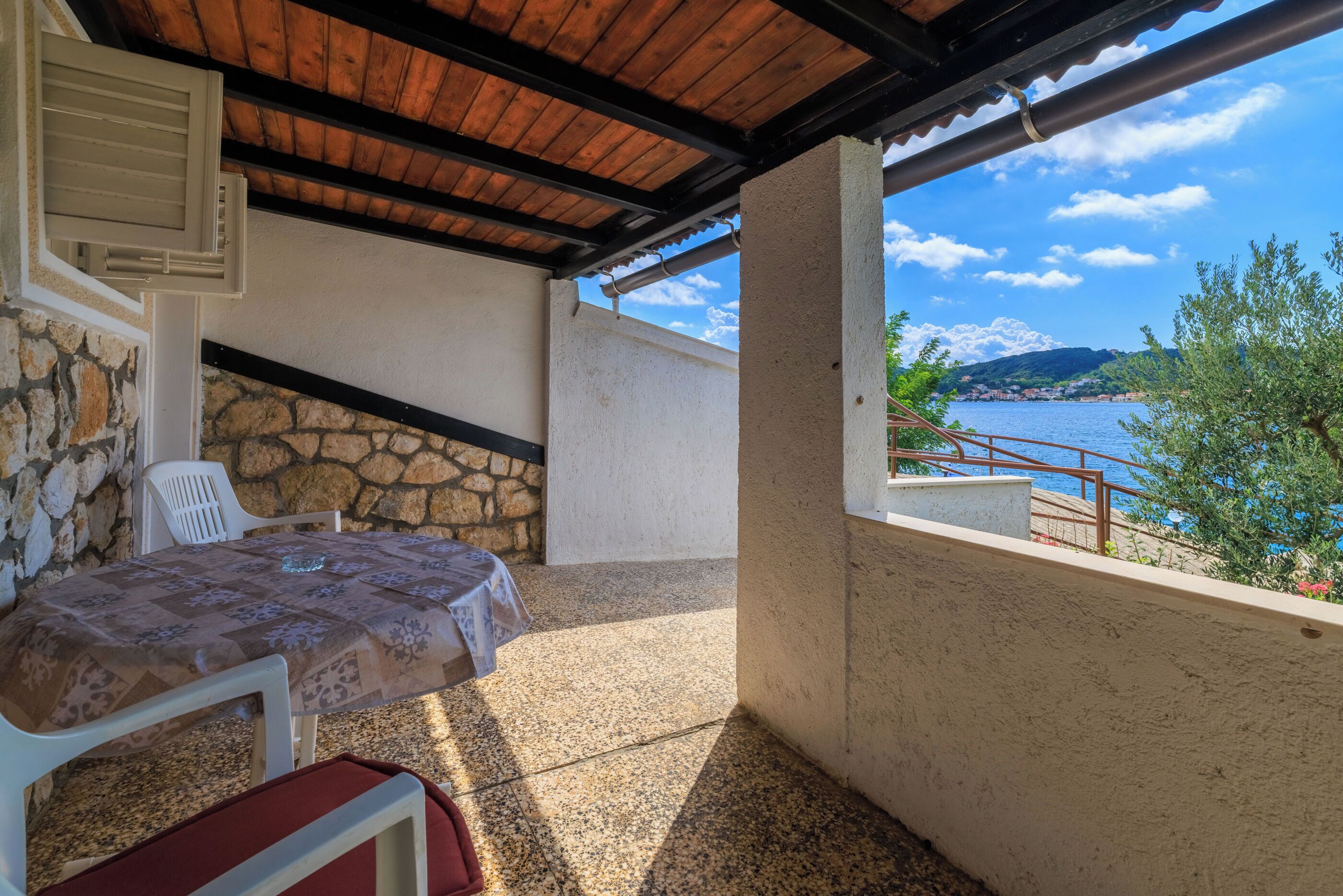 Un espace extérieur aménagé, abrité, avec une table et des chaises, un mur en pierre et une vue sur une étendue d’eau et des collines.