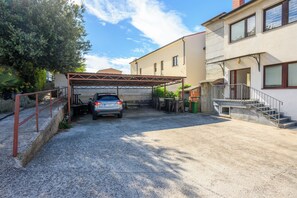 Un espace de stationnement couvert avec une voiture, un bâtiment doté d’un escalier et un jardin.