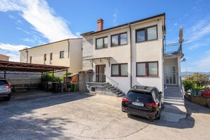 Un edificio de dos pisos con un porche cubierto, un coche estacionado al frente y vista al mar de fondo.