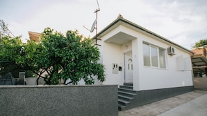 A small, single-story building with a white exterior and a grey base. There is a small tree in front of the building, and a satellite dish is mounted on the roof. - Apartment Florio (Trogir)