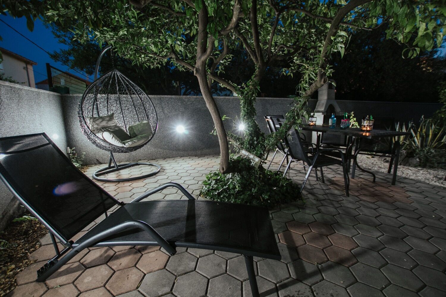 An outdoor patio with hexagonal tiles, a hanging chair, a table with chairs, and a small garden area.