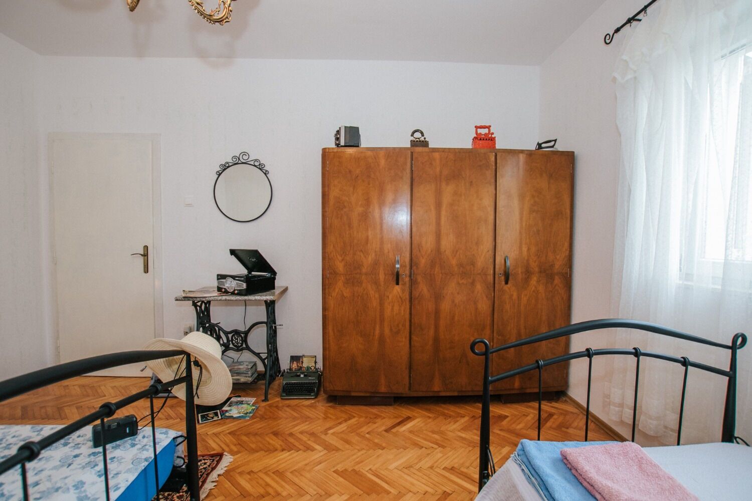 A bedroom with a wooden wardrobe, a bed with a metal frame, a small table with a radio, and a mirror on the wall.
