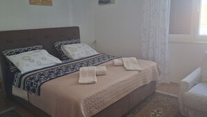 Un lit avec une couvre-lit à motifs, des oreillers et une serviette pliée. Il y a une fenêtre avec des rideaux et une chaise sur la droite.