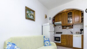 Un espace de vie compact comprenant un canapé, un petit coin cuisine avec un réfrigérateur, un four et des placards en bois, ainsi qu’un tableau accroché au mur.