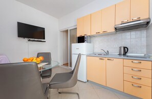 A compact kitchen with a microwave, stove, and sink. There is a dining table with chairs and a bowl of fruit. A flat-screen TV is mounted on the wall. - Apartment Karlo (Dugi Rat)