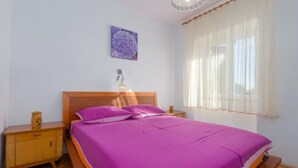 A bedroom with a wooden bed, purple bedding, a nightstand with a lamp, and a window with sheer curtains. - Apartment Lili (Umag)