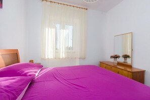 Ein Schlafzimmer mit einem Bett, einer Holzkommode, einem Spiegel und einem Fenster mit durchsichtigen Vorhängen.
