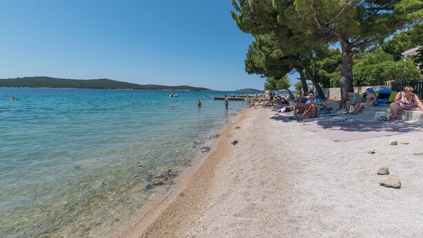 Une plage à l’eau claire, où l’on peut voir des gens se prélasser au soleil, et une zone bordée d’arbres.