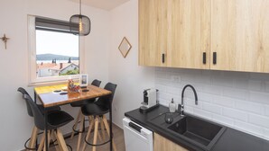 Une cuisine compacte avec une table en bois, des chaises noires, un lave-vaisselle encastré, un évier noir et une fenêtre donnant sur des bâtiments et des collines.