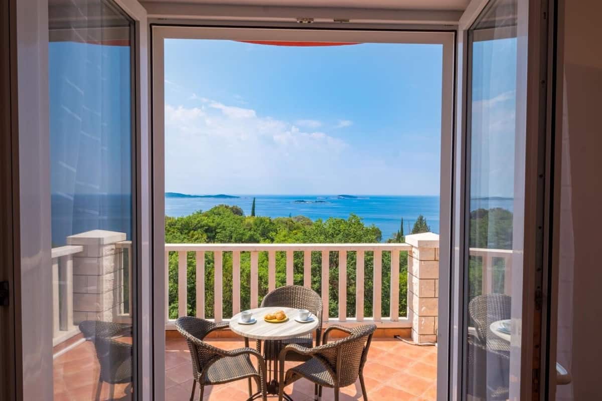 A balcony with a table and chairs, overlooking a scenic view of the sea and greenery.