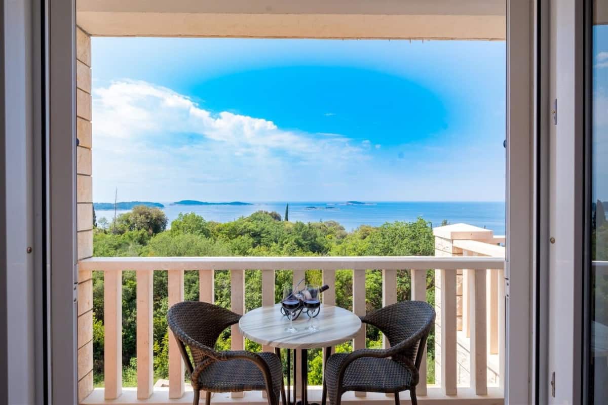 A balcony with wicker chairs and a table, overlooking a scenic view of the sea and greenery.