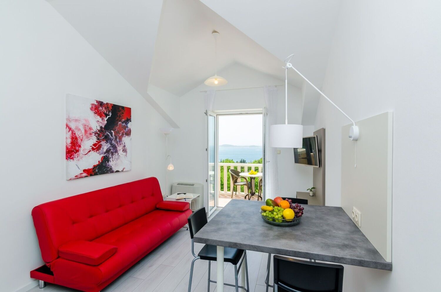 A modern living room with a red sofa, a dining table with fruit, and a balcony with a view.