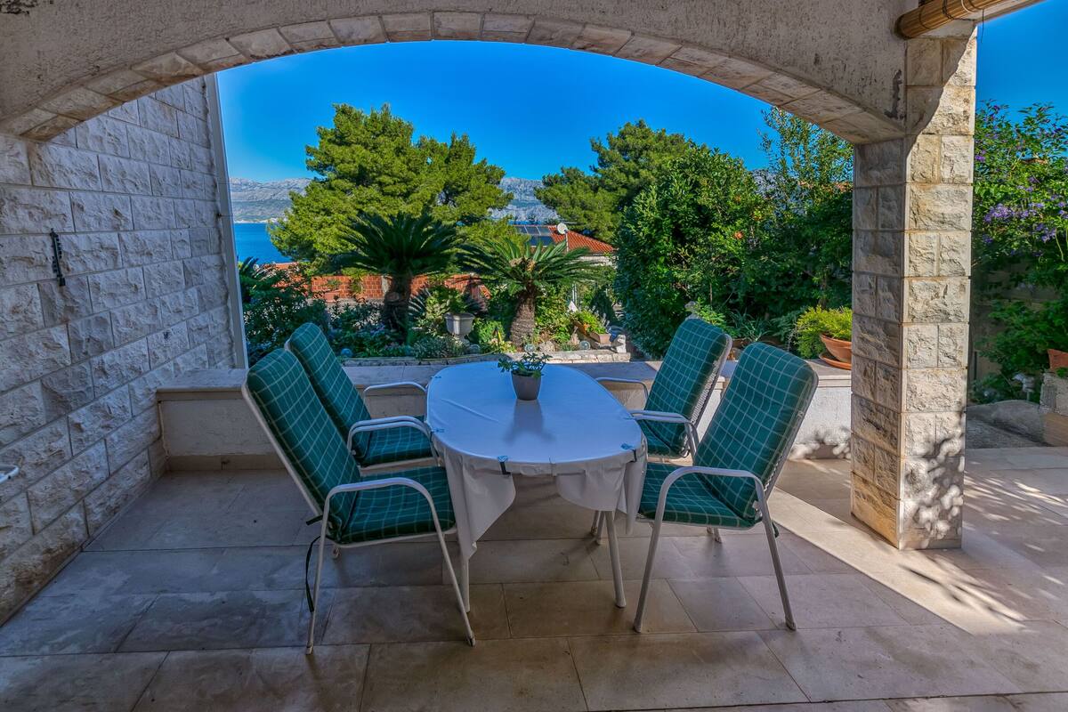 A stone archway leading to a patio with a round table and four chairs, overlooking a scenic view of trees and a body of water.