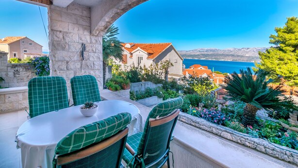 A terrace with a stone wall, a table with chairs, and a view of the sea and houses. - Apartments Paco (Postira)