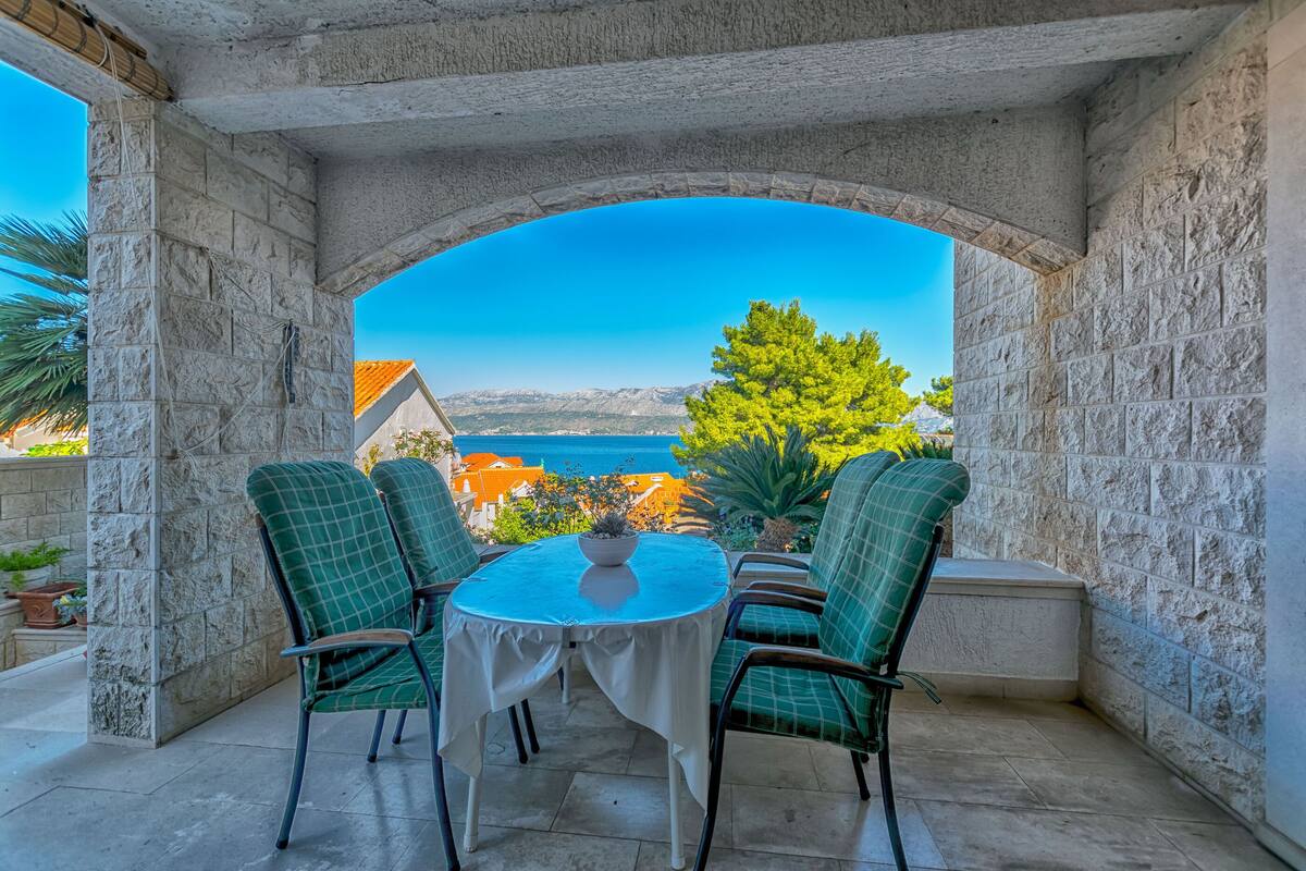 A stone-arched outdoor seating area with a table and chairs, overlooking a scenic view of a coastal town and mountains.