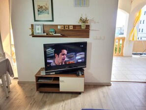 A living room with a flat-screen TV on a wooden stand, a shelf with decorative items, and a balcony with yellow curtains. - Apartments Mare Family Only (Kolan)