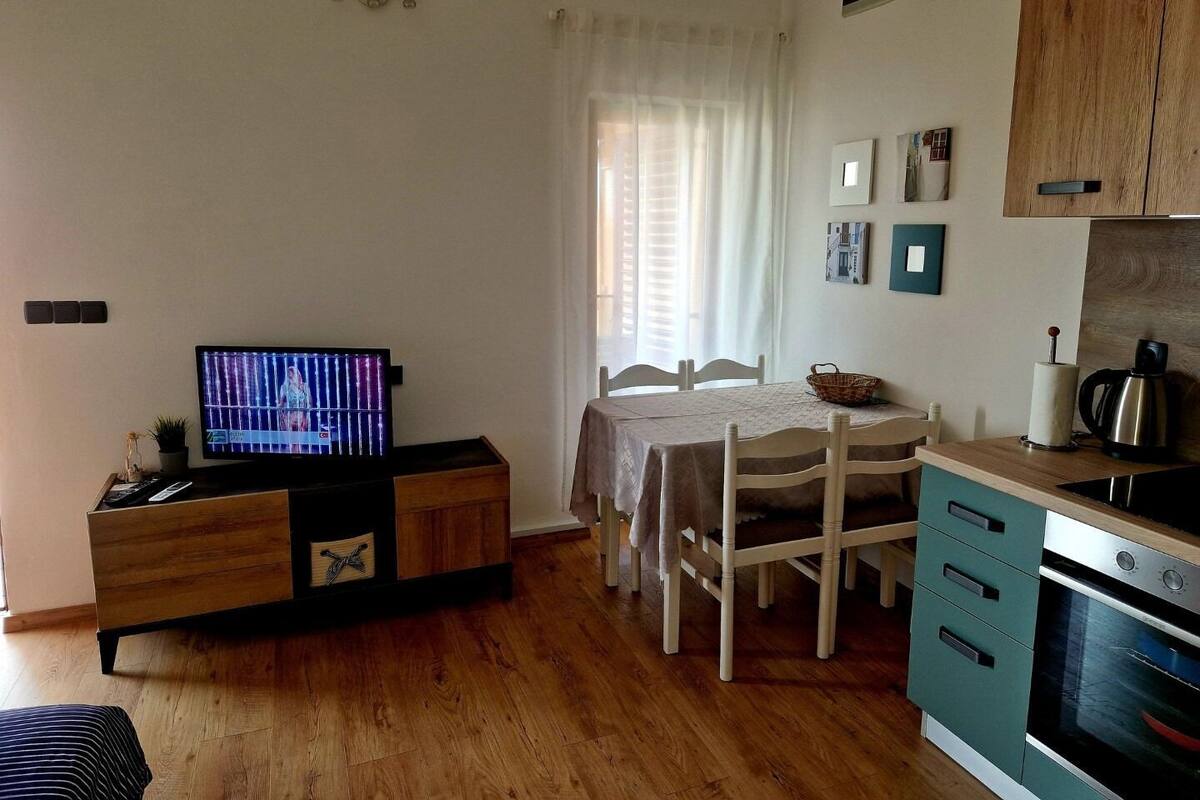 A compact living space with a wooden dining table, a television, a kitchenette with a stove, and a window with curtains.