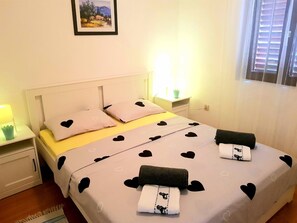 A bedroom with a bed, two bedside tables, a nightstand, a window with blinds, and a framed picture on the wall. - Apartments Mare Family Only (Kolan)