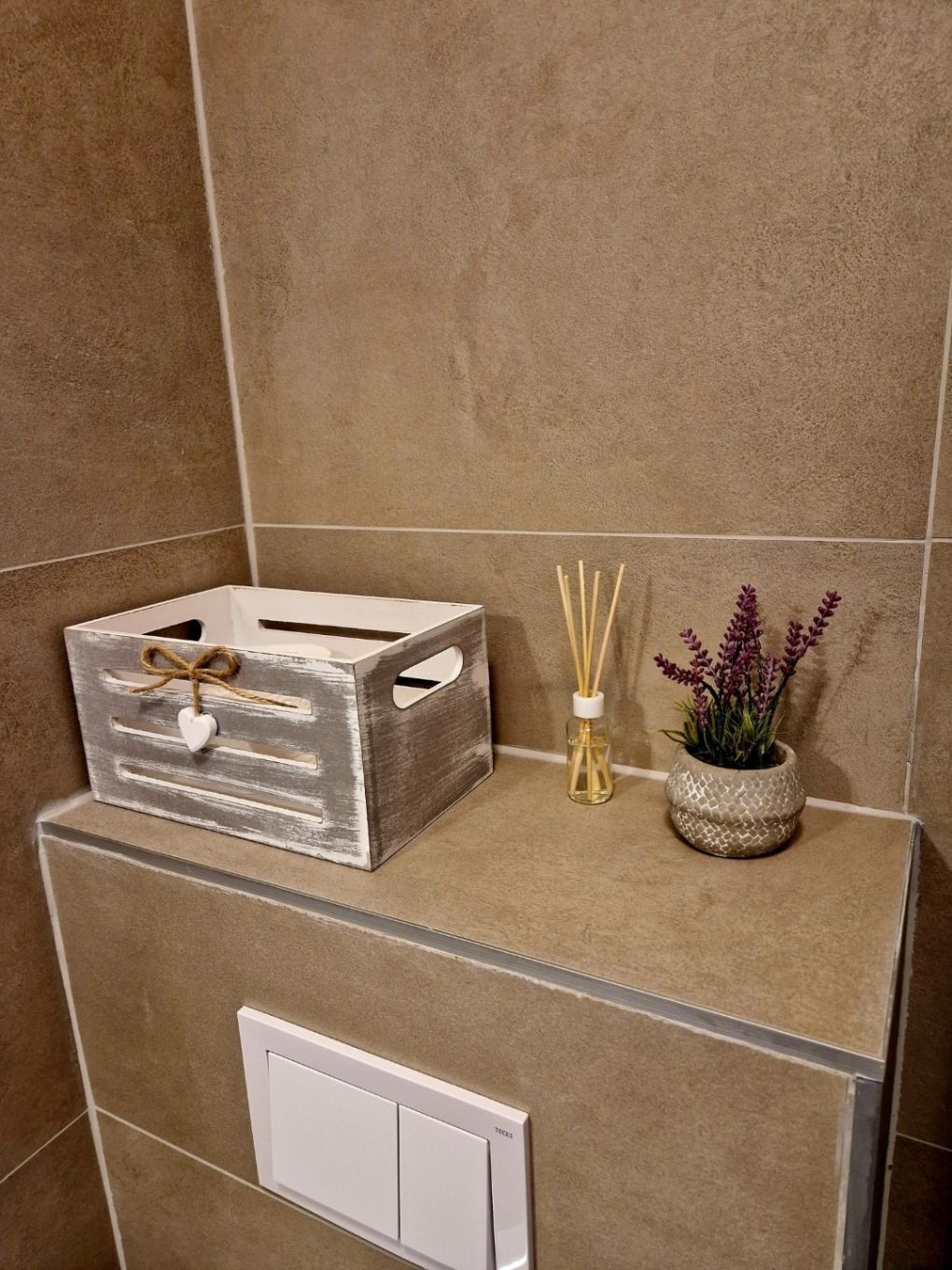 Um banheiro com porta-toalhas branco, caixa de madeira com decoração de coração, difusores de vime e uma pequena planta em vaso.