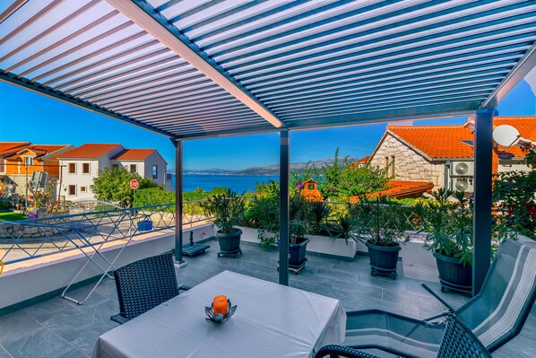 A covered outdoor seating area with a table and chairs, potted plants, and a view of the sea and buildings in the background. - Apartments Terrace (Postira)