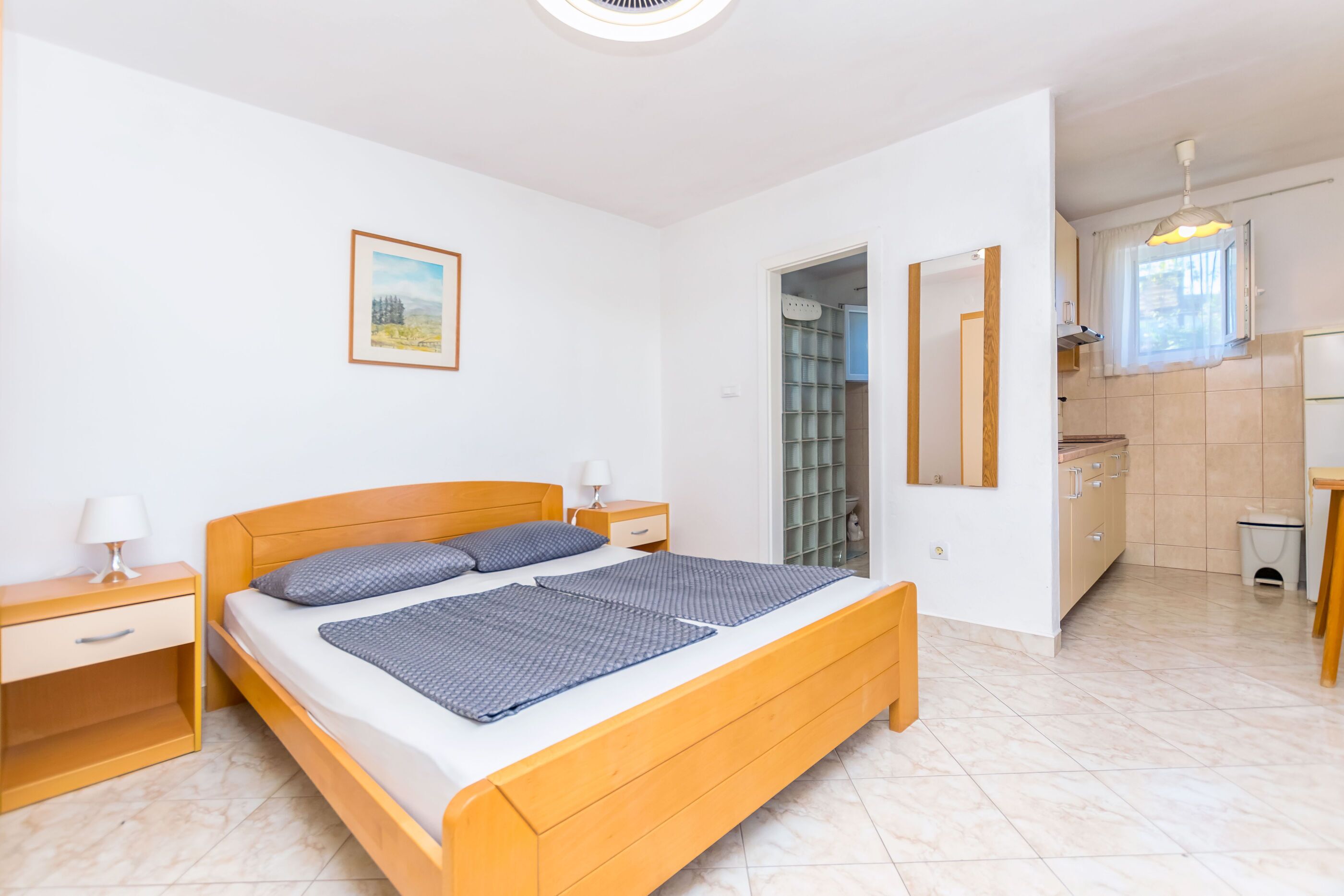 Un dormitorio con cama de madera, mesitas de noche, un tocador y un cuadro en la pared, además de un baño visible a través de una puerta abierta.