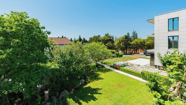 A modern building with a flat roof, surrounded by a well-maintained garden with various trees and shrubs. - Apartments Ivo (Kastela)