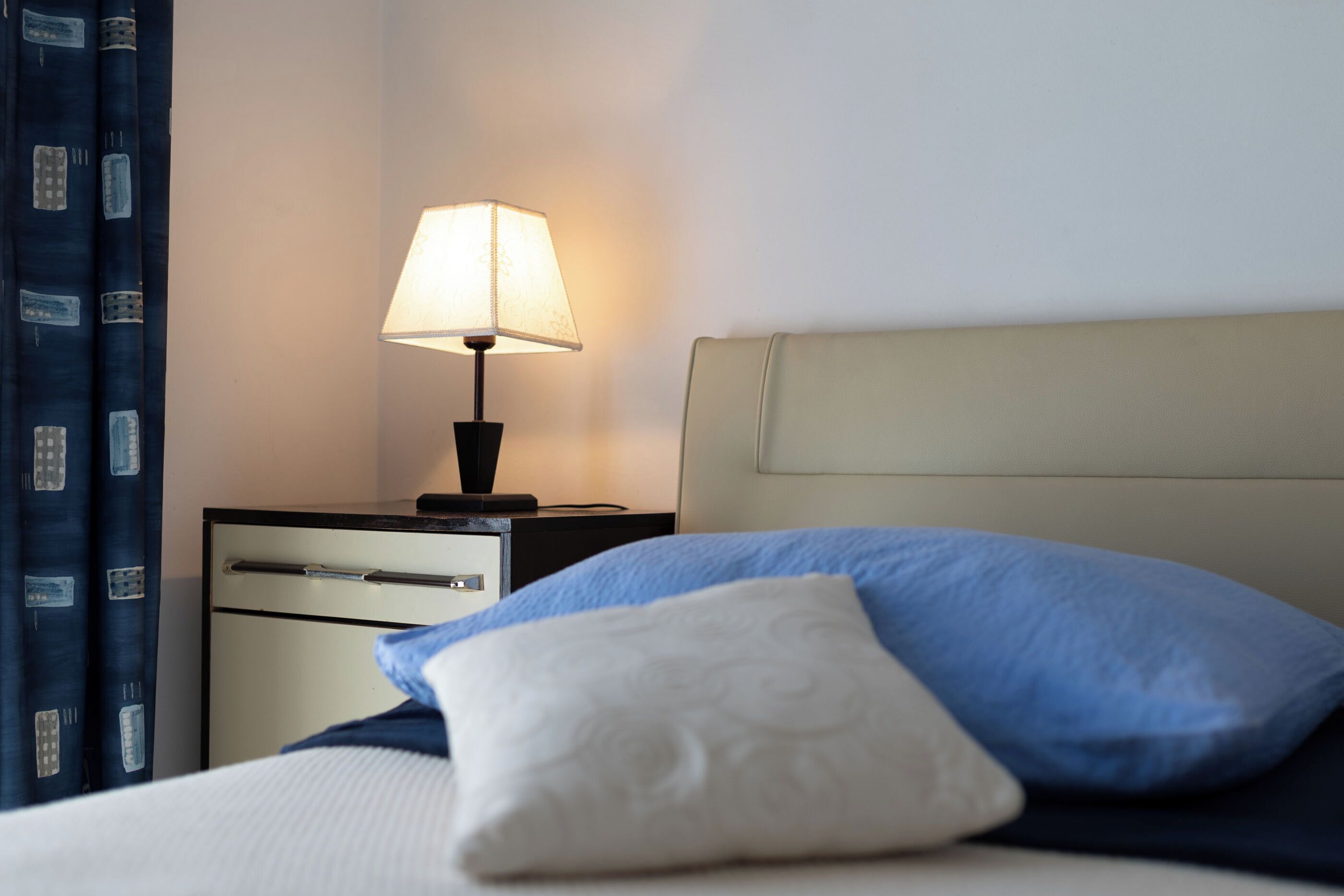 A hotel room with a bed, pillows, a nightstand, a lamp, and a curtain.