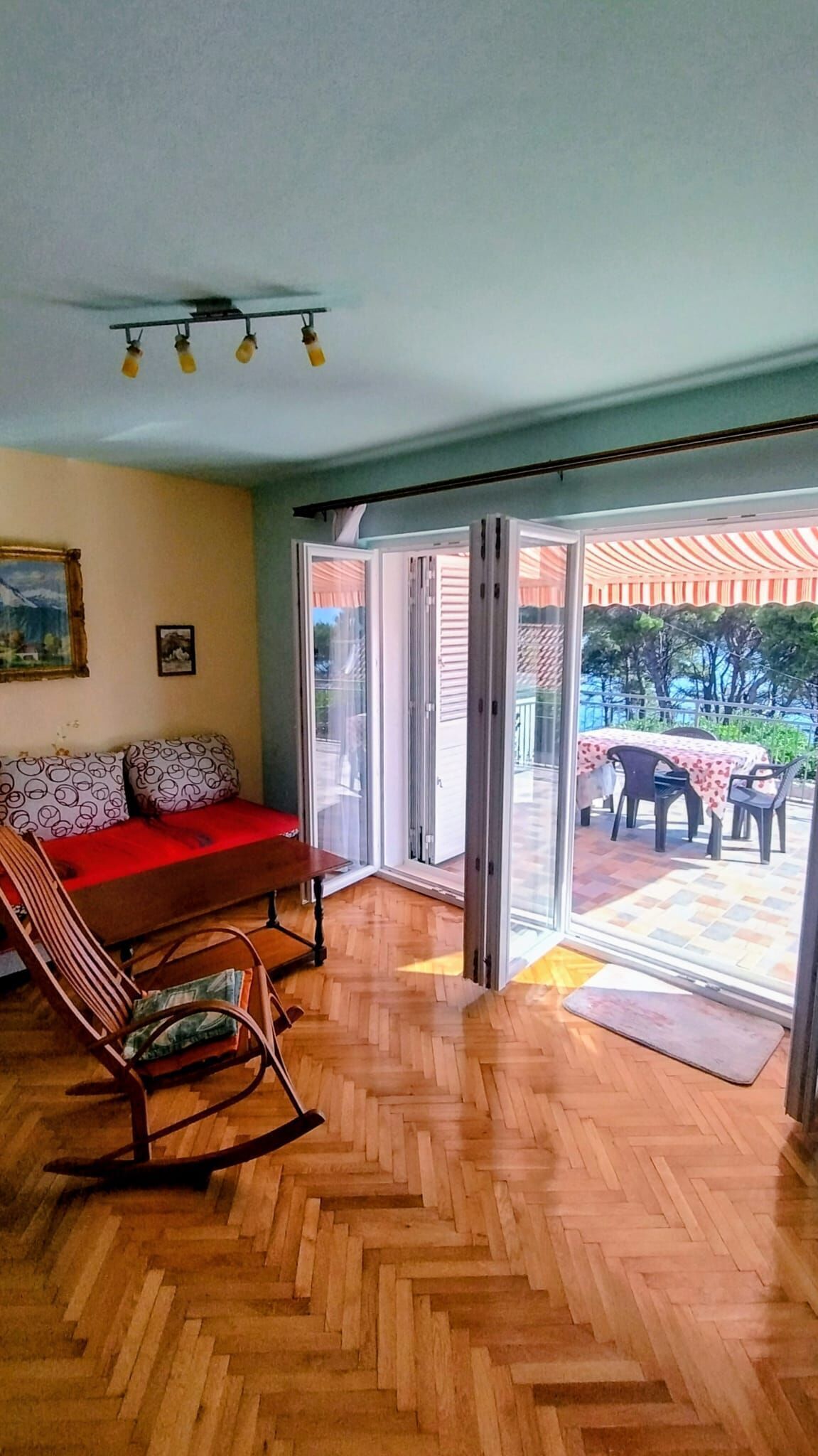 A room with a wooden herringbone floor, a red sofa, a rocking chair, and a sliding glass door leading to an outdoor patio with a table and chairs.