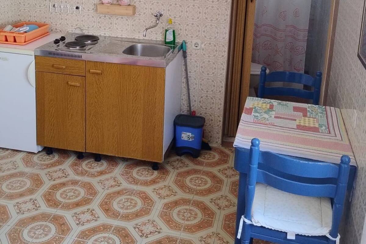 A compact kitchen with a stove, sink, and wooden cabinets. There is a small table with a chair, a blue trash bin, and a clock on the wall.