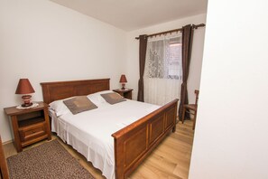 A bedroom with a wooden bed, two bedside tables, a nightstand, a chair, and a window with curtains. - Apartment Grdelin (Biograd na Moru)