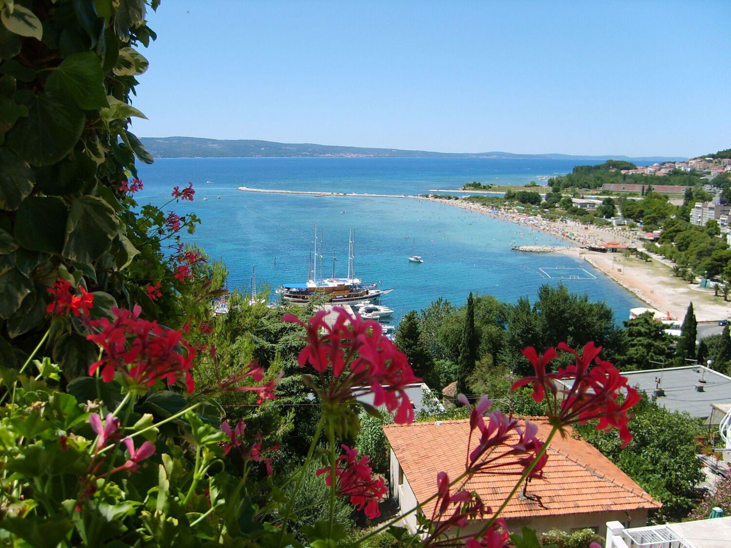 A coastal town with boats docked at a marina, surrounded by greenery and buildings with red-tiled roofs.