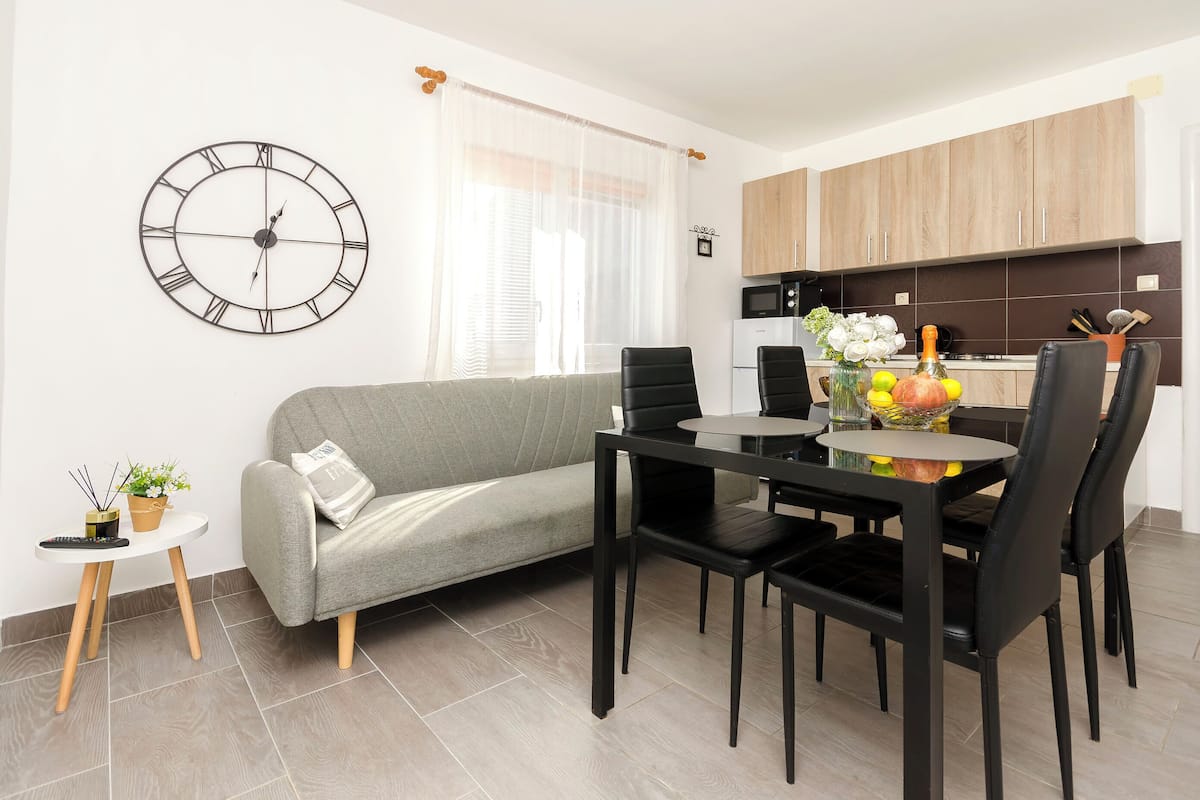A modern living space with a sofa, dining table, chairs, a clock, and a kitchen area.