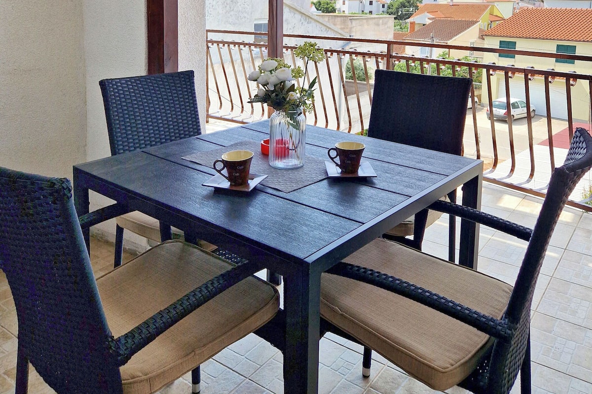 A balcony with a table set for two, chairs, and a vase with flowers.