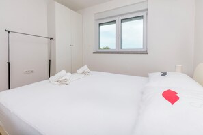 A neatly made bed with white linens, a window with a view, and a black metal rack against the wall. - Apartment Cvita (Primosten)