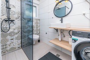 A modern bathroom with patterned tiles, a glass shower enclosure, a toilet, a sink with a mirror, and a washing machine. - Apartment Cvita (Primosten)
