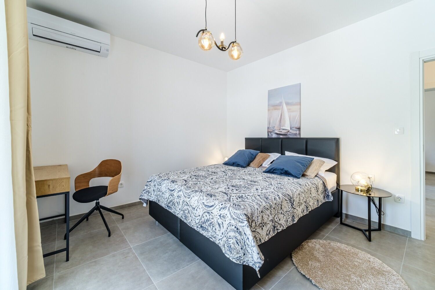 A bedroom with a bed, a desk, a chair, and a bedside table with a lamp.
