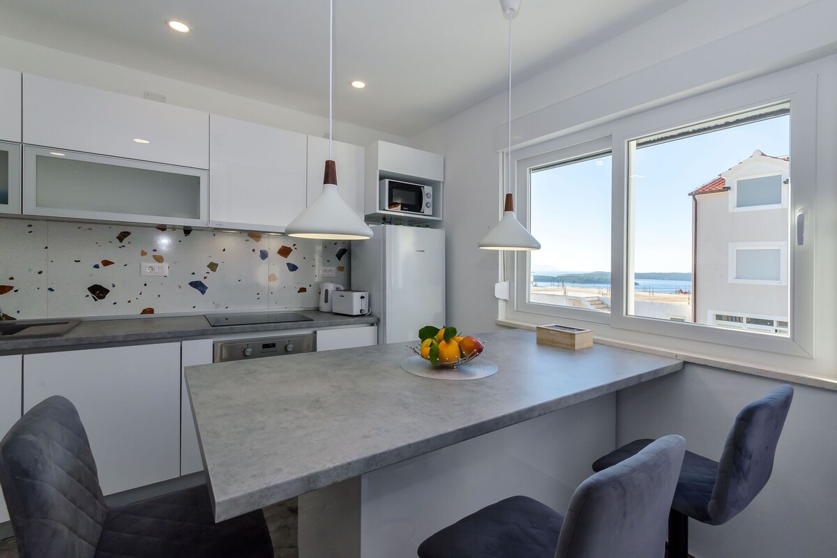 A modern kitchen with white cabinets, a granite countertop, and a view of the beach through large windows.