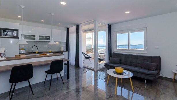 Une cuisine moderne dotée d’un îlot central, d’un coin repas avec des chaises, d’un canapé et d’une vue sur un balcon extérieur, accessible par une porte vitrée.