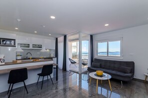 Een moderne keuken met een centraal eiland, een eethoek met stoelen, een bank en uitzicht door een glazen deur op een buitenbalkon.