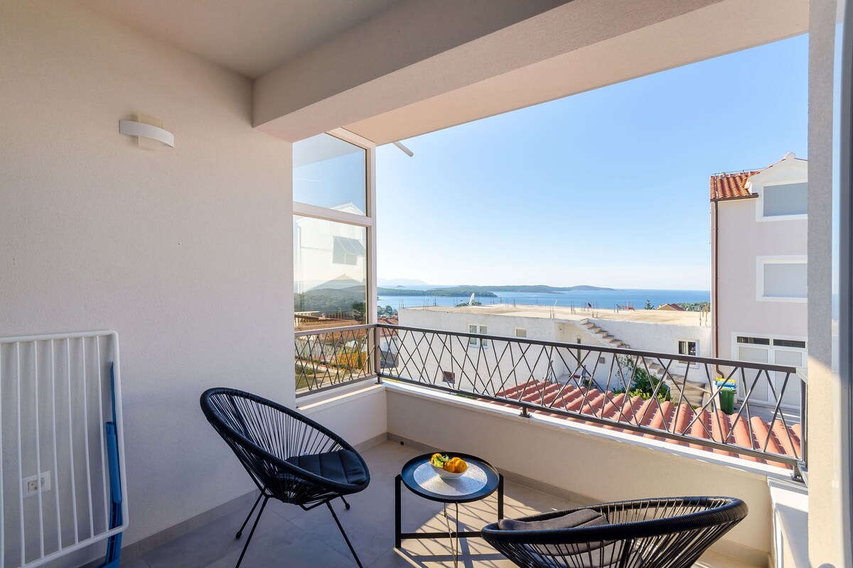 A balcony with a view of the sea, a small table with fruit, and two chairs.