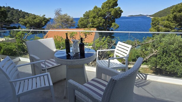 Un balcon agrémenté de meubles en osier, une table ronde avec une bouteille et un verre, et une vue sur la mer et les collines.