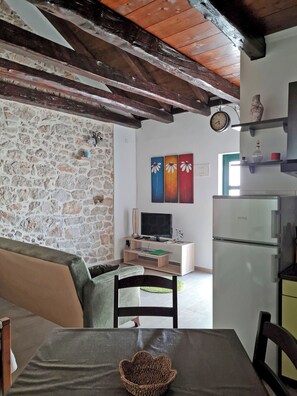 A room with a stone wall, a wooden ceiling, a television on a wooden stand, a refrigerator, and a dining table with a basket. - Apartments Studio Zaton (Nin)