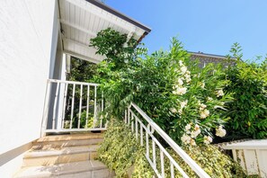A staircase leading to a white building with a white railing, surrounded by lush greenery and flowering plants. - Apartments Rosuje (Omisalj)