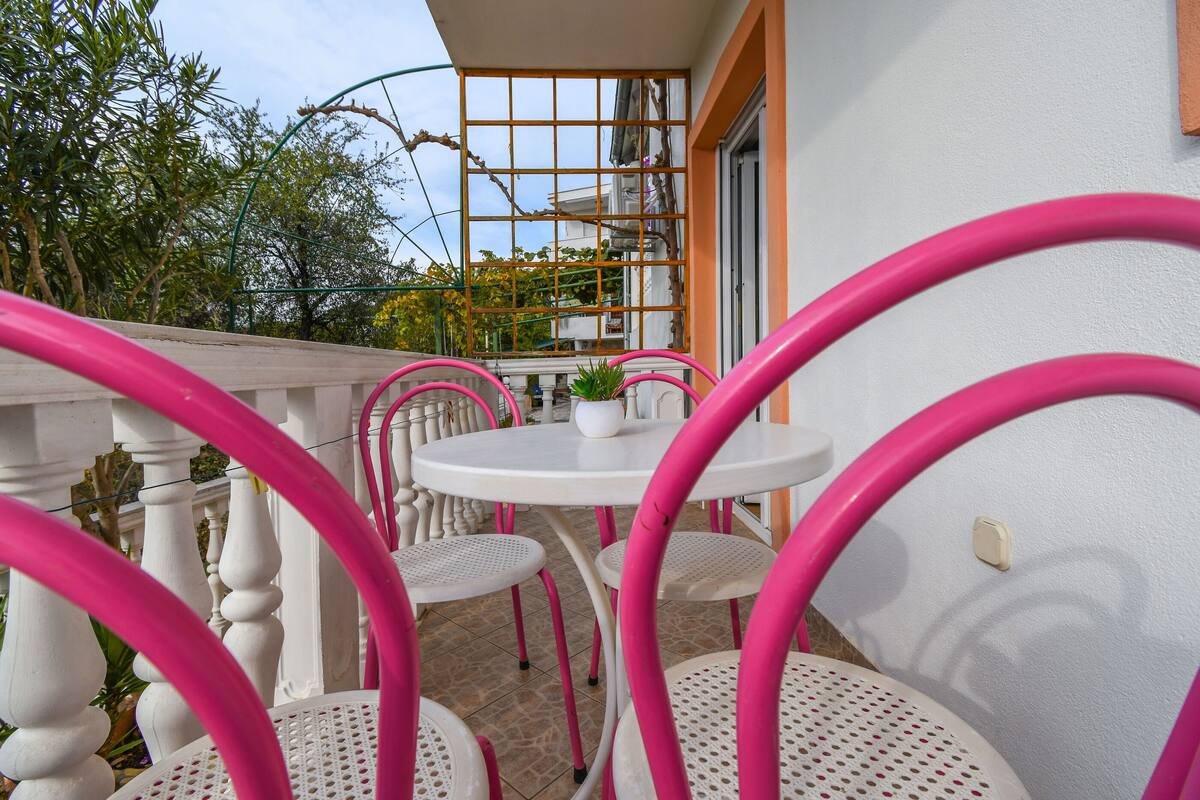 A balcony with white chairs and a table, a window with a view of trees and buildings, and a railing.