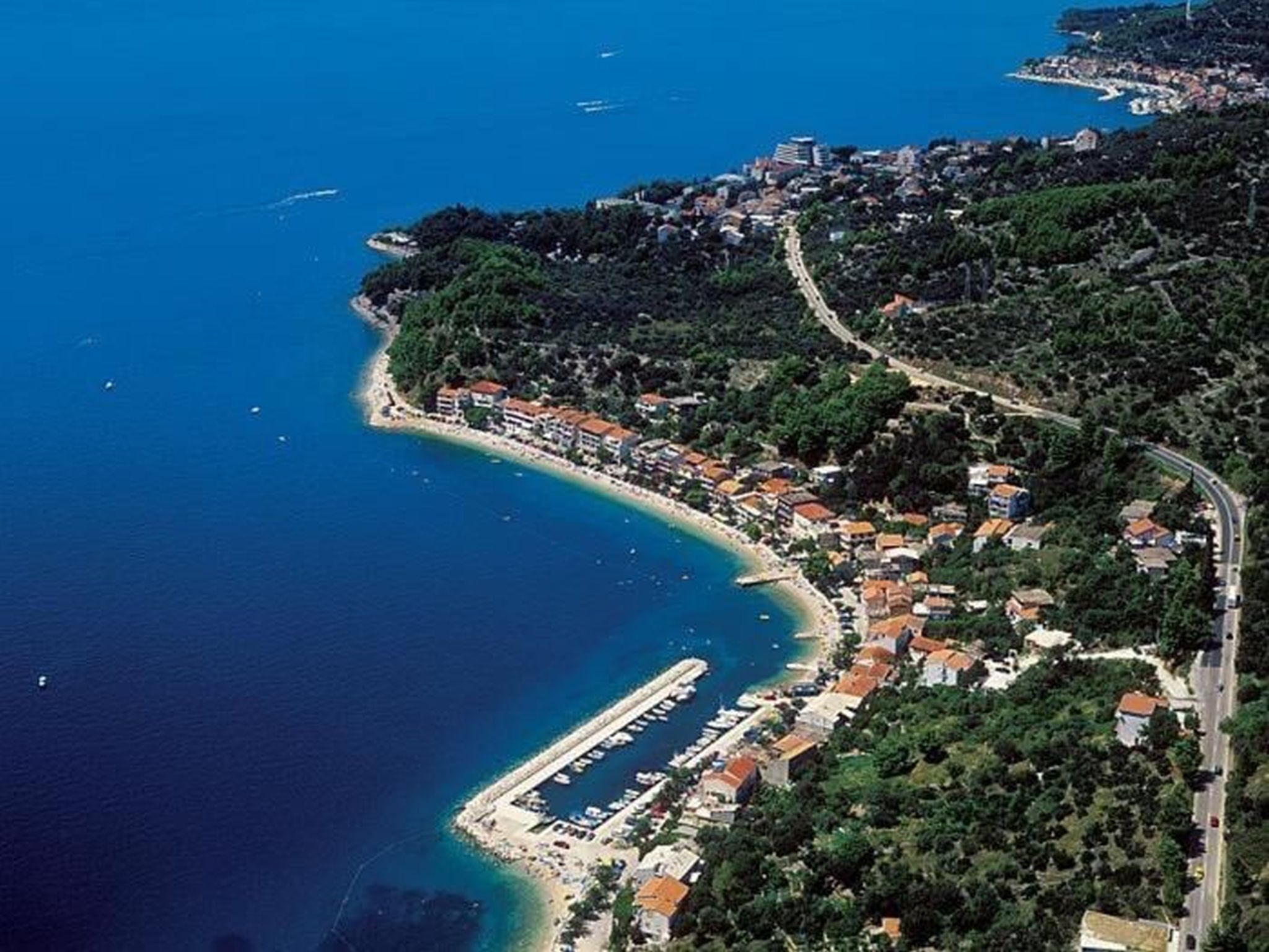 An aerial view of a coastal town with a marina, dense greenery, and a winding road.